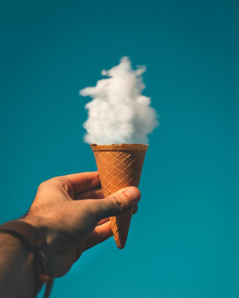 Hand holding an ice cream cone with a cloud effect against a clear sky background.